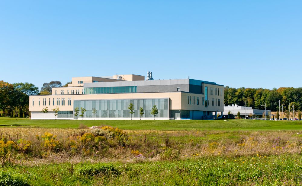 A view of the new Lakehead University building showing the rurual-like surroundings of grassy fields, and mature trees.